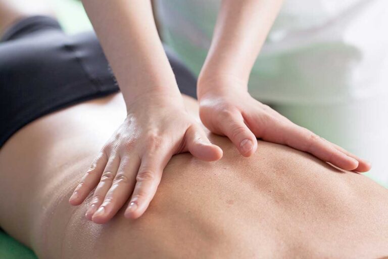 A chiropractor gently adjusting the upper back of an adult in a bright, sunlit treatment room.